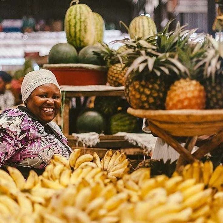 A smiling female fruit vendor at Kimironko Market in Kigali, Rwanda, surrounded by vibrant displays of yellow bananas, pineapples, and watermelons. Photo by Roadrave Tours and Travel.