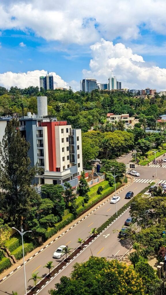High-angle view of a clean, modern street in Kigali, Rwanda, featuring a divided road with palm trees, contemporary apartment buildings, and the city skyline under a blue sky with white clouds.